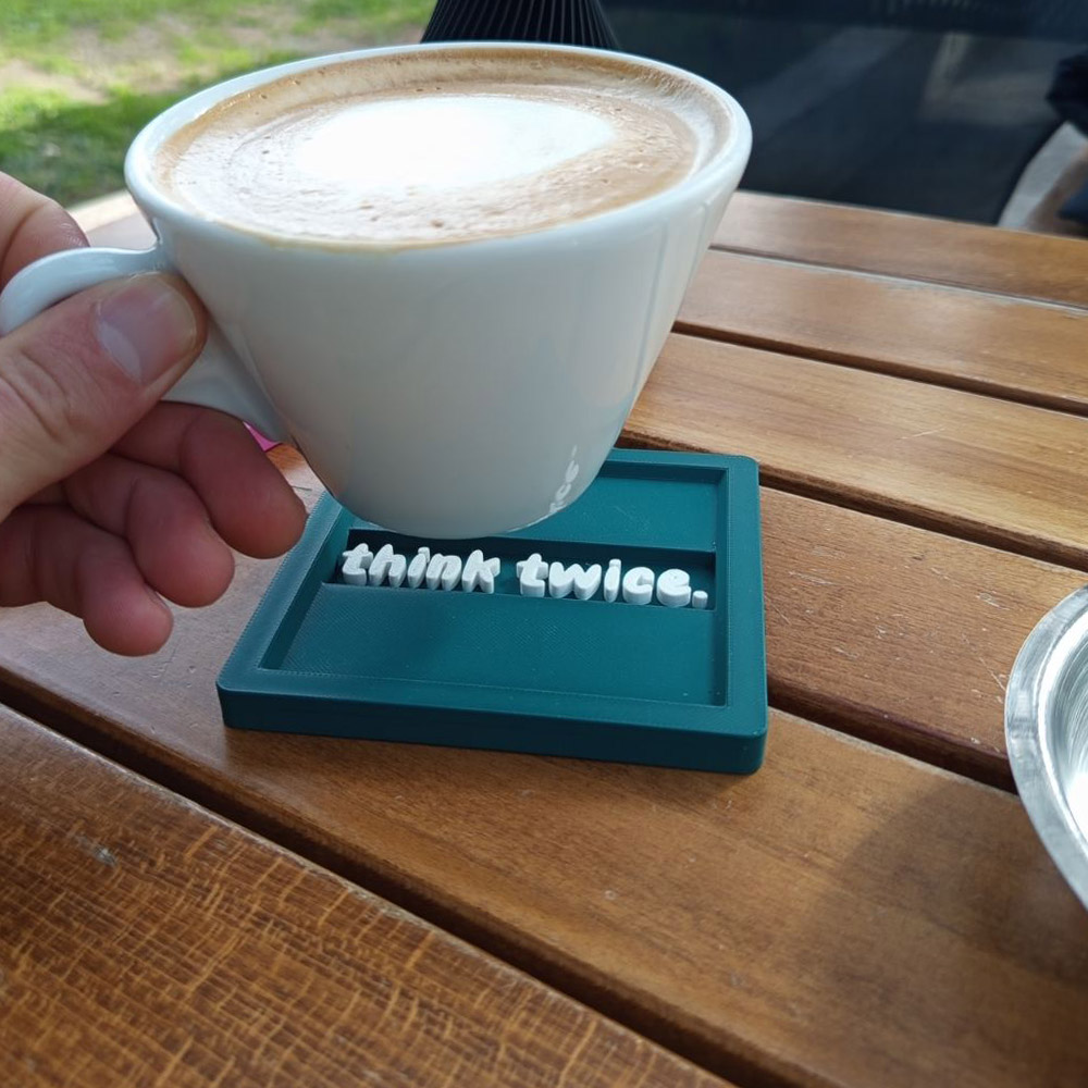 3D printed σουβέρ “think twice.” σε πράσινο Morandi με λευκά ανάγλυφα γράμματα, με κούπα καφέ τοποθετημένη επάνω, σε ξύλινο τραπέζι.